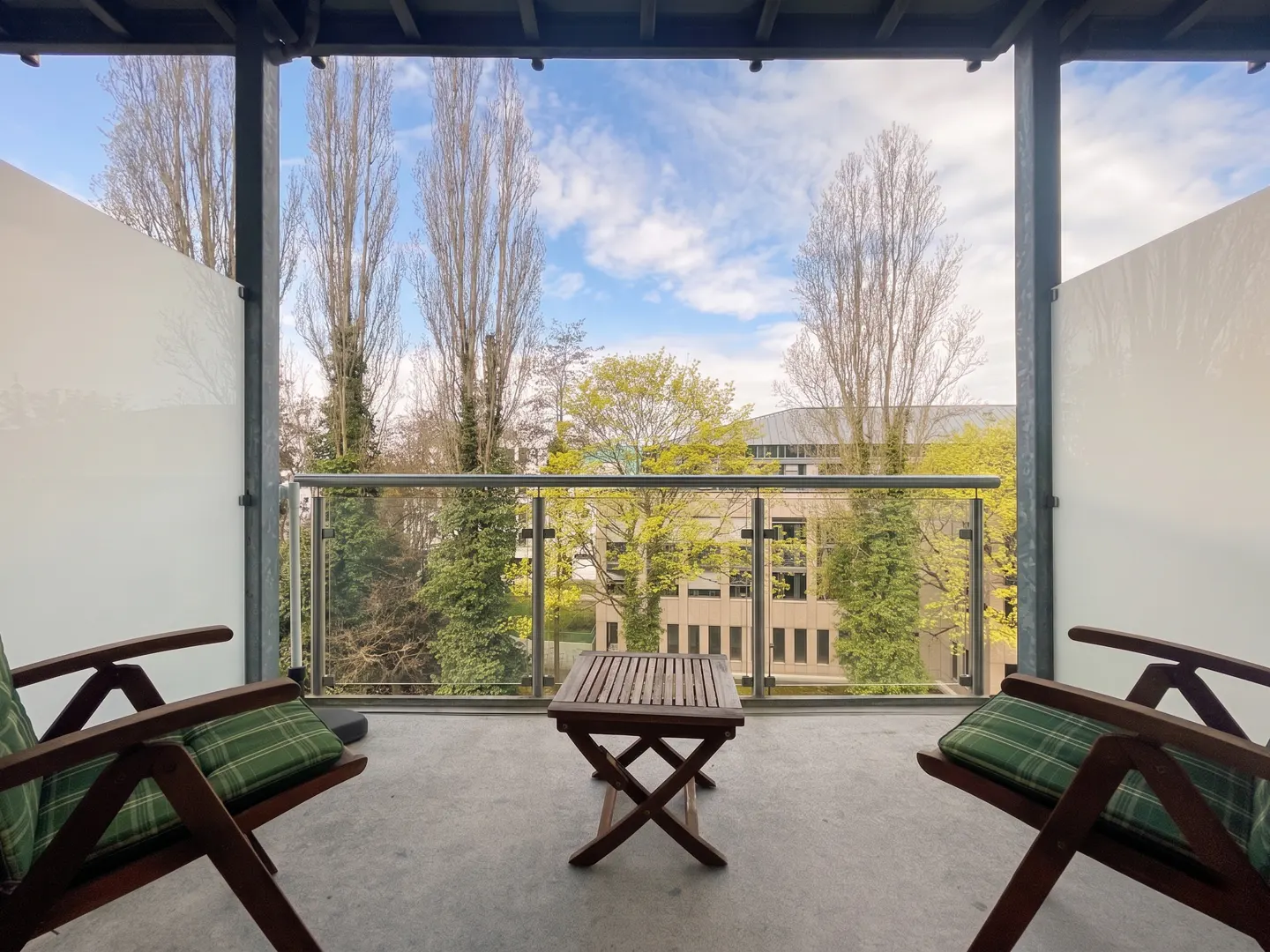 Balcony view with two wooden chairs, green plaid cushions, and a small wooden table. Trees and a building are visible beyond the glass railing.