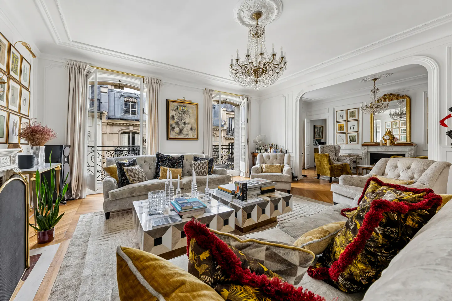 Bright living room with sofas, chairs, and a crystal chandelier. Balconies overlook a European city.