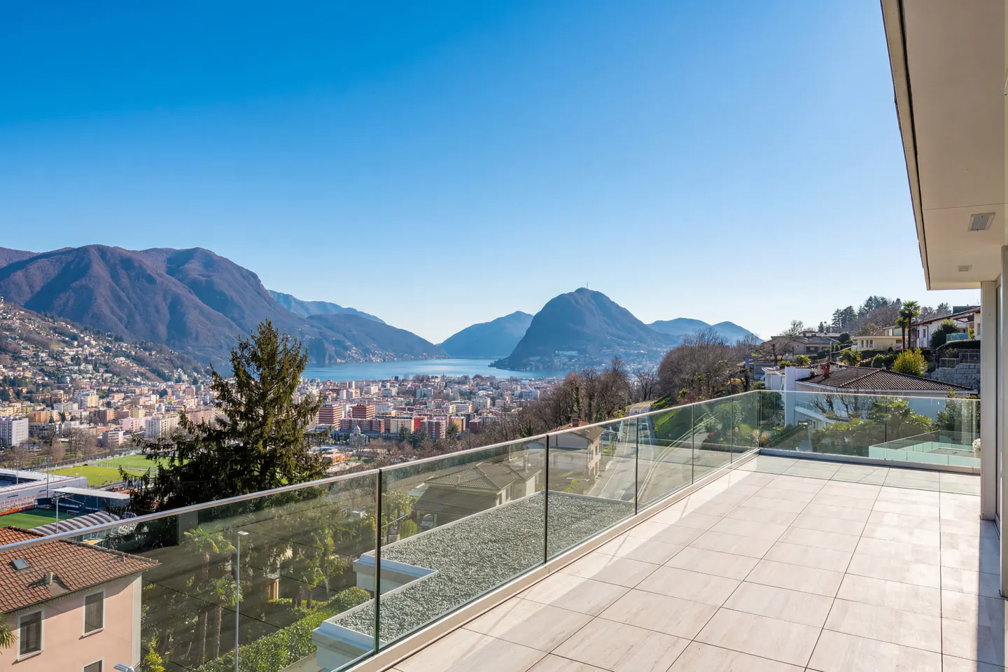 View from a modern balcony with glass railings overlooking a city, lake, and mountains under a clear blue sky.