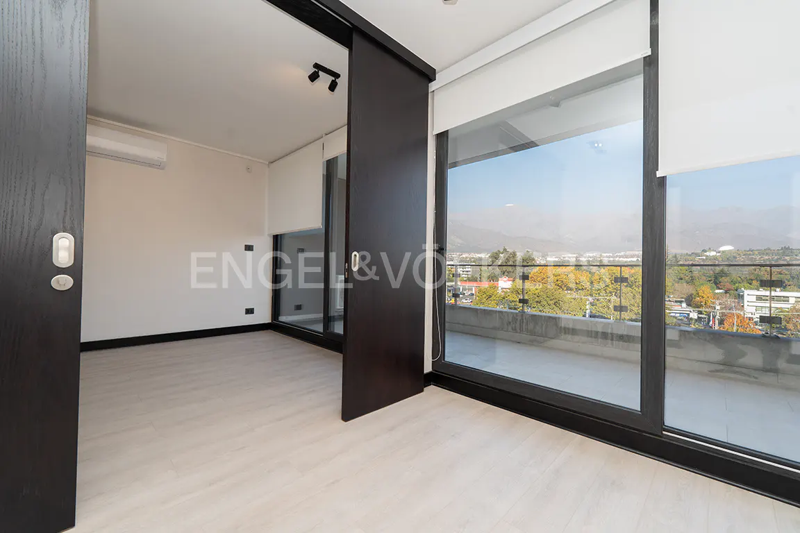 A bright, empty room with light wood floors and a sliding glass door to a balcony with a city view.