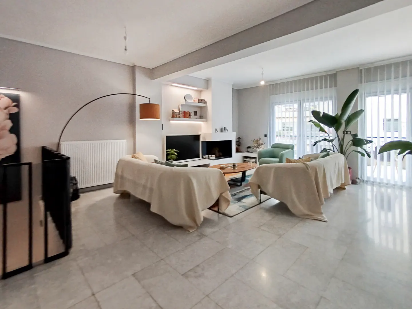 Bright living room with marble floors, two sofas draped in beige throws, a fireplace, and a large plant near the window.