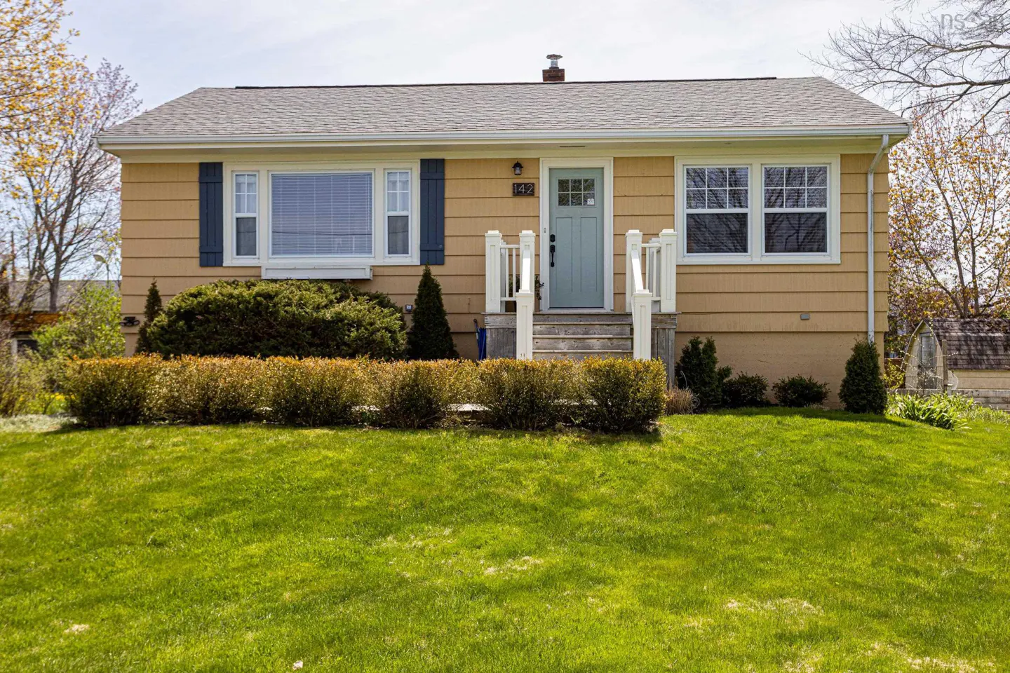 A tan ranch-style house with a light blue door and dark blue shutters. A green lawn and trimmed bushes are in front.