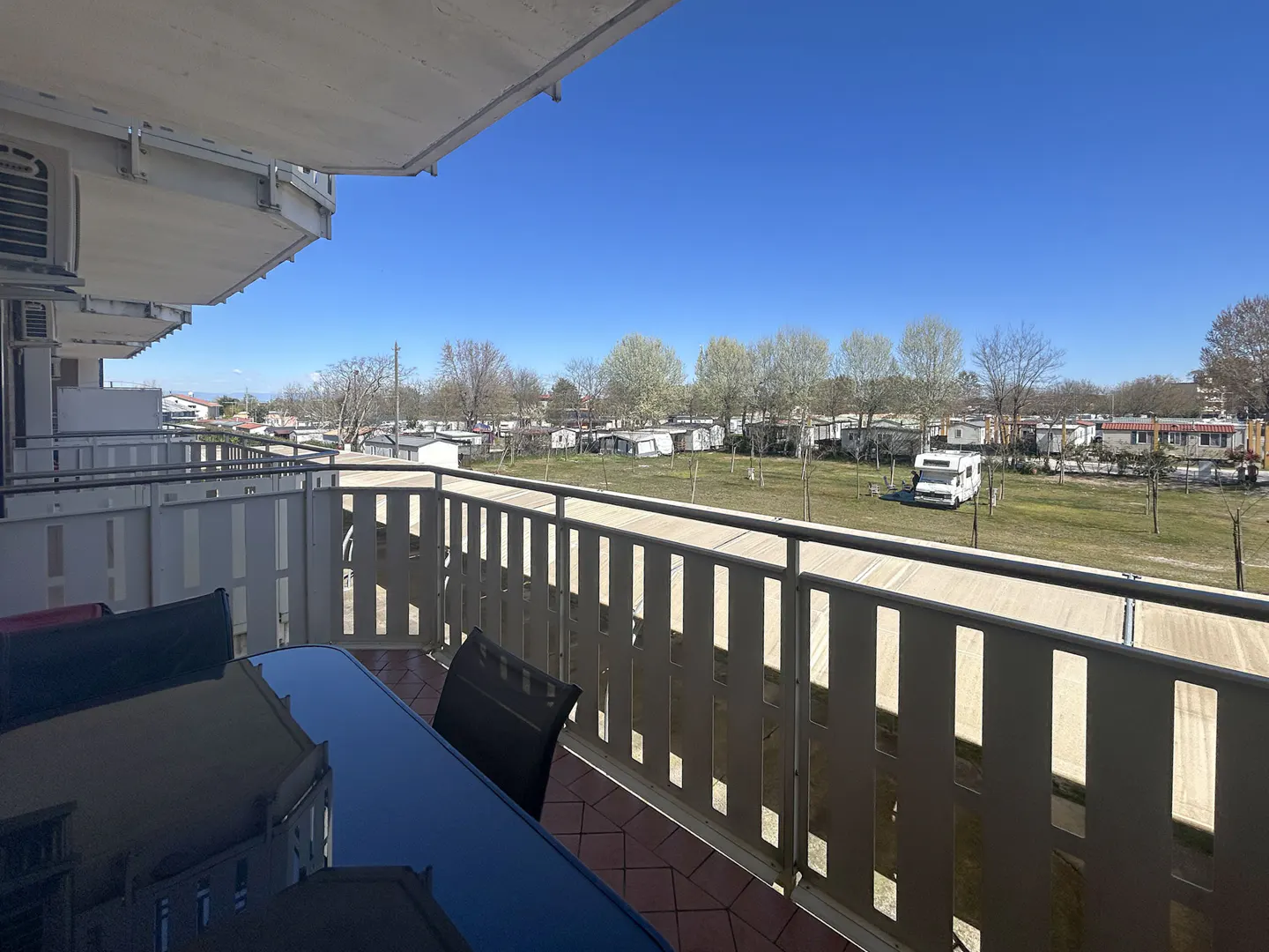 Balcony view of a sunny campground. White railings overlook RVs and trees under a clear blue sky. A dark table and chairs sit on the tiled balcony.