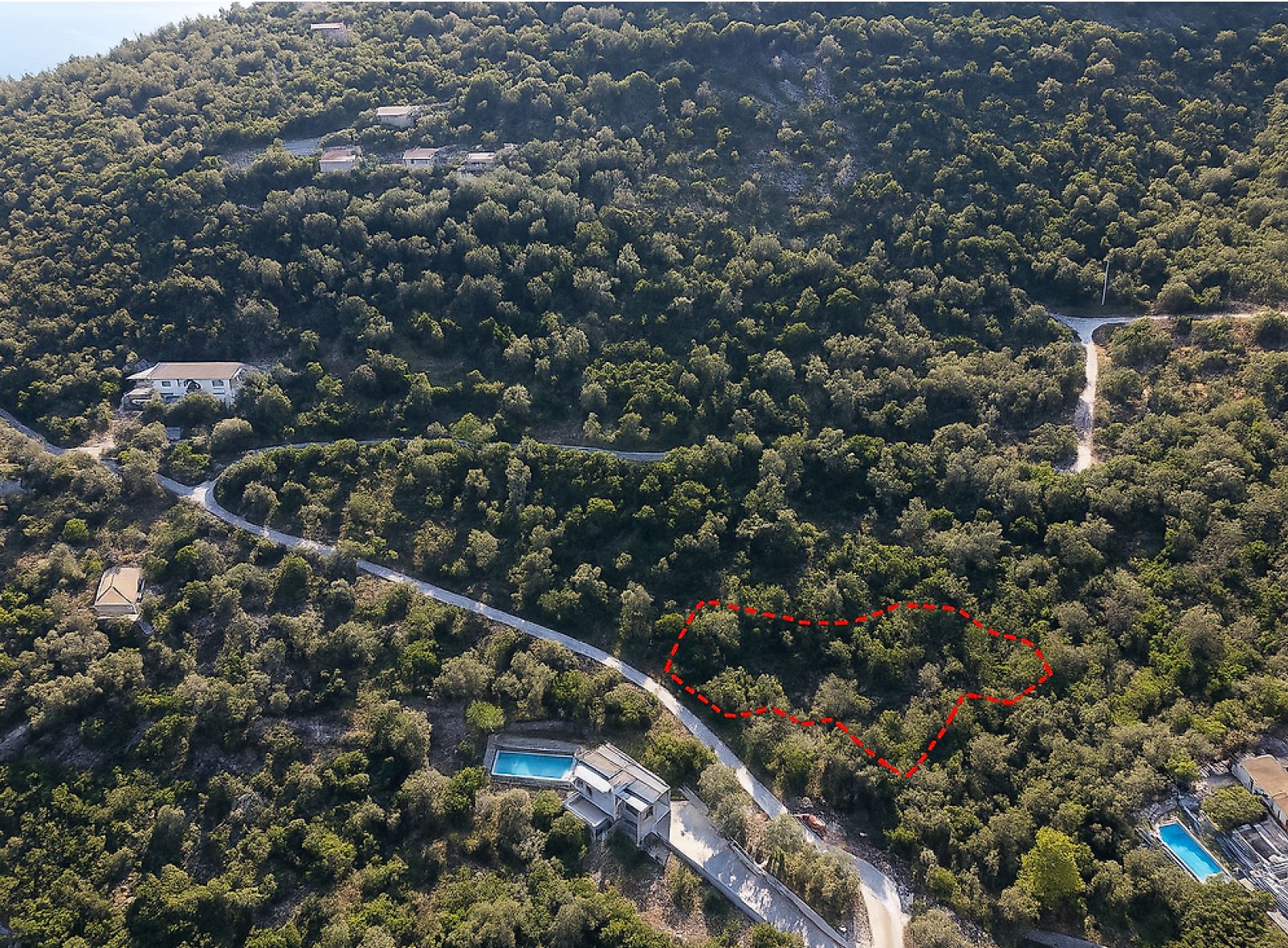 Aerial view of a wooded hillside with a winding road, scattered houses, and a red dashed outline marking a plot of land.