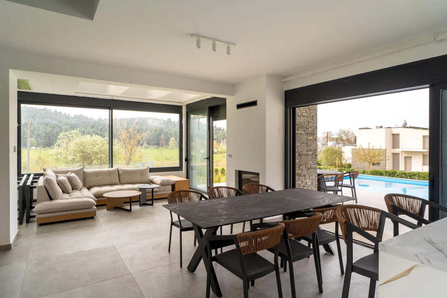 Bright, modern living room with a beige sectional sofa, black dining table and chairs, and a view of a pool and green landscape.