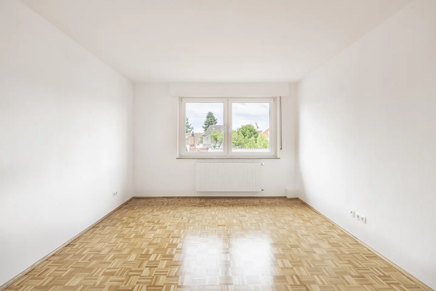 Empty room with white walls, parquet floor, and a window showing trees and houses outside. A white radiator is under the window.