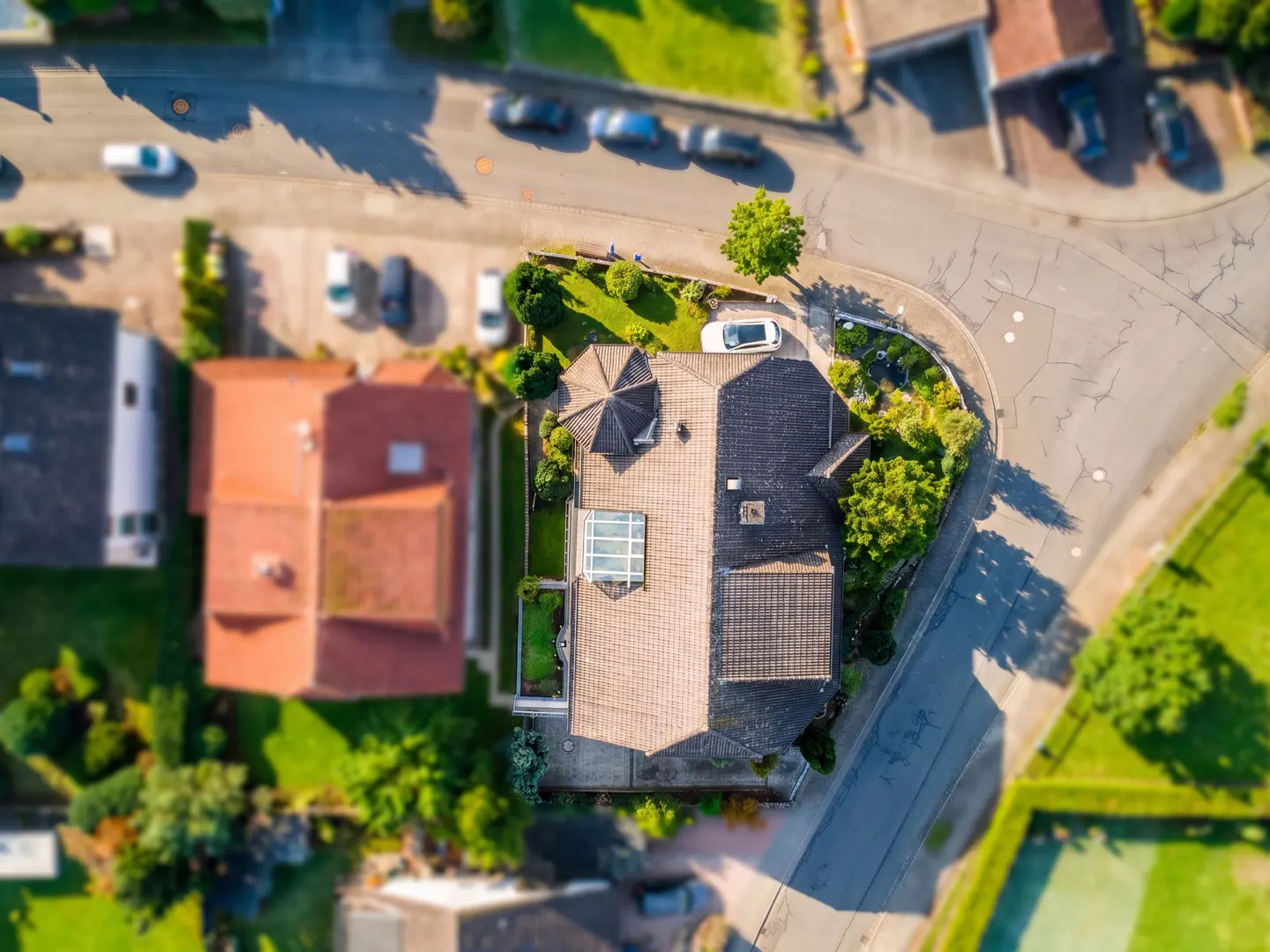 Aerial view of a house with a gray roof, green lawn, and a white car parked in the driveway. Cars are parked on the street.