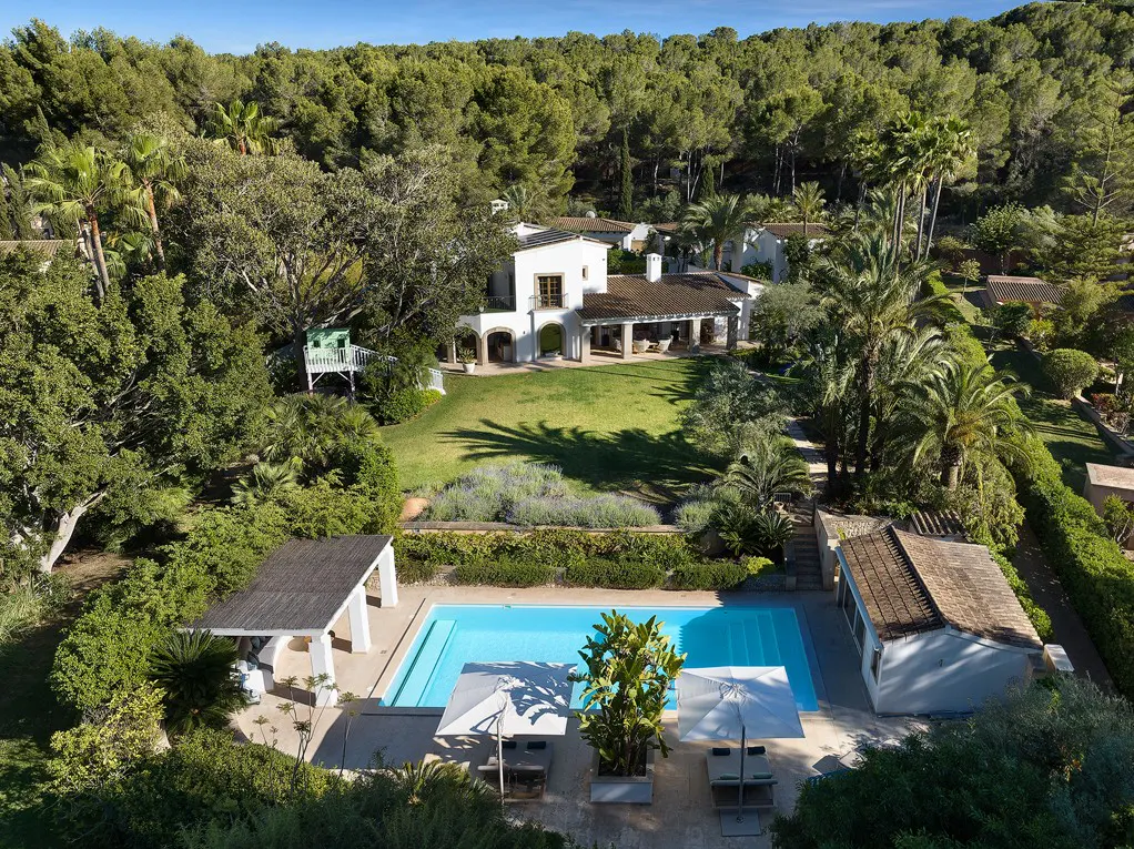 Aerial view of a white house with a pool, surrounded by lush greenery and trees. Umbrellas and lounge chairs are near the pool.