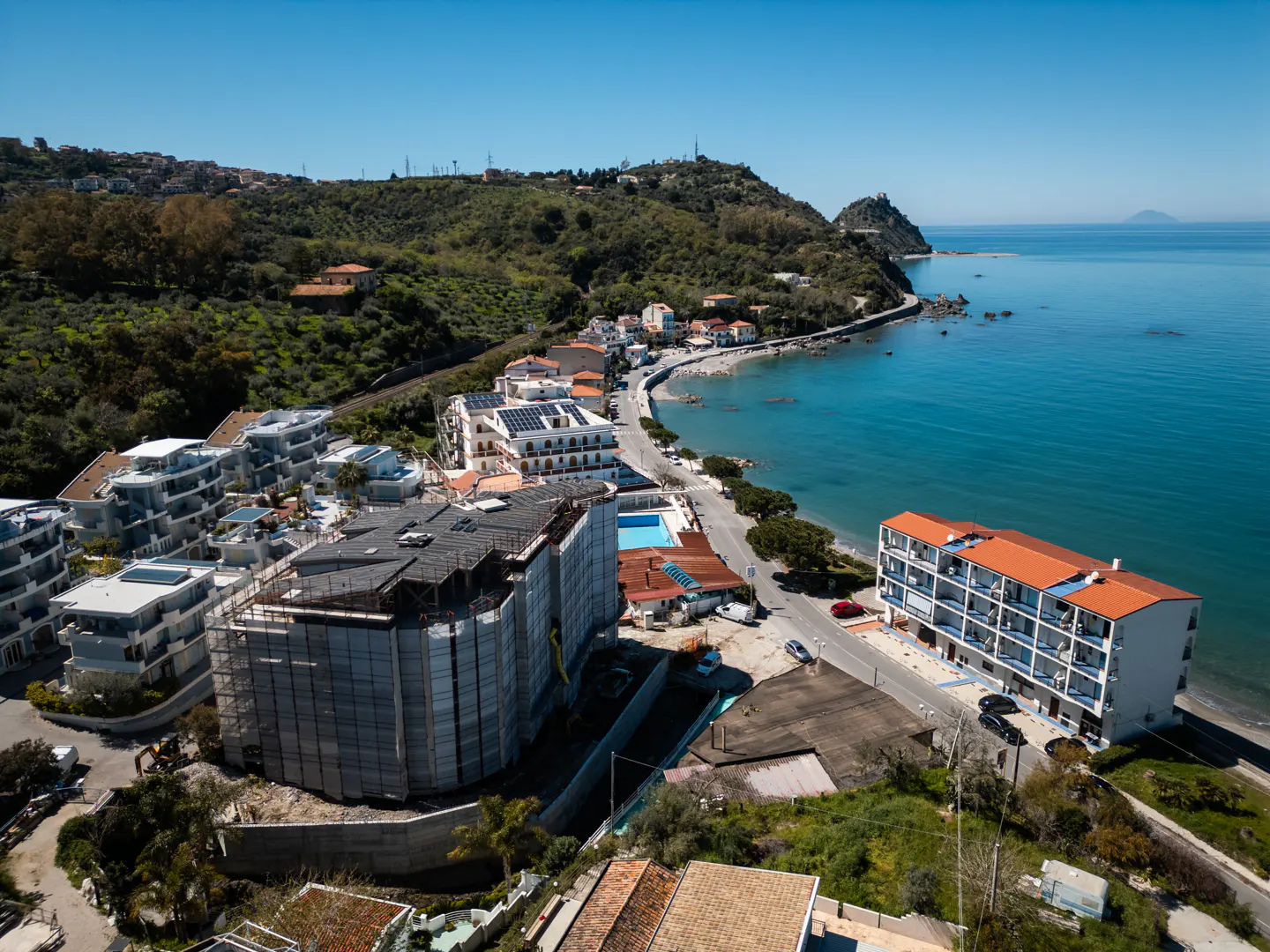 Coastal view of buildings and blue sea. New construction is wrapped in gray material. Hills are green and the sky is blue.