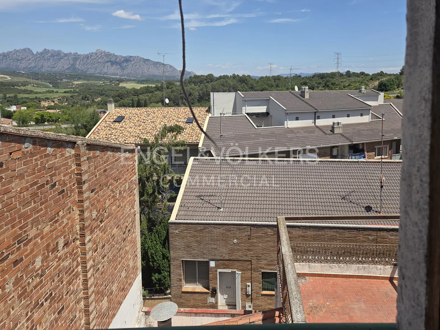 View from a balcony overlooking a residential neighborhood with tiled roofs and a distant mountain range under a blue sky.