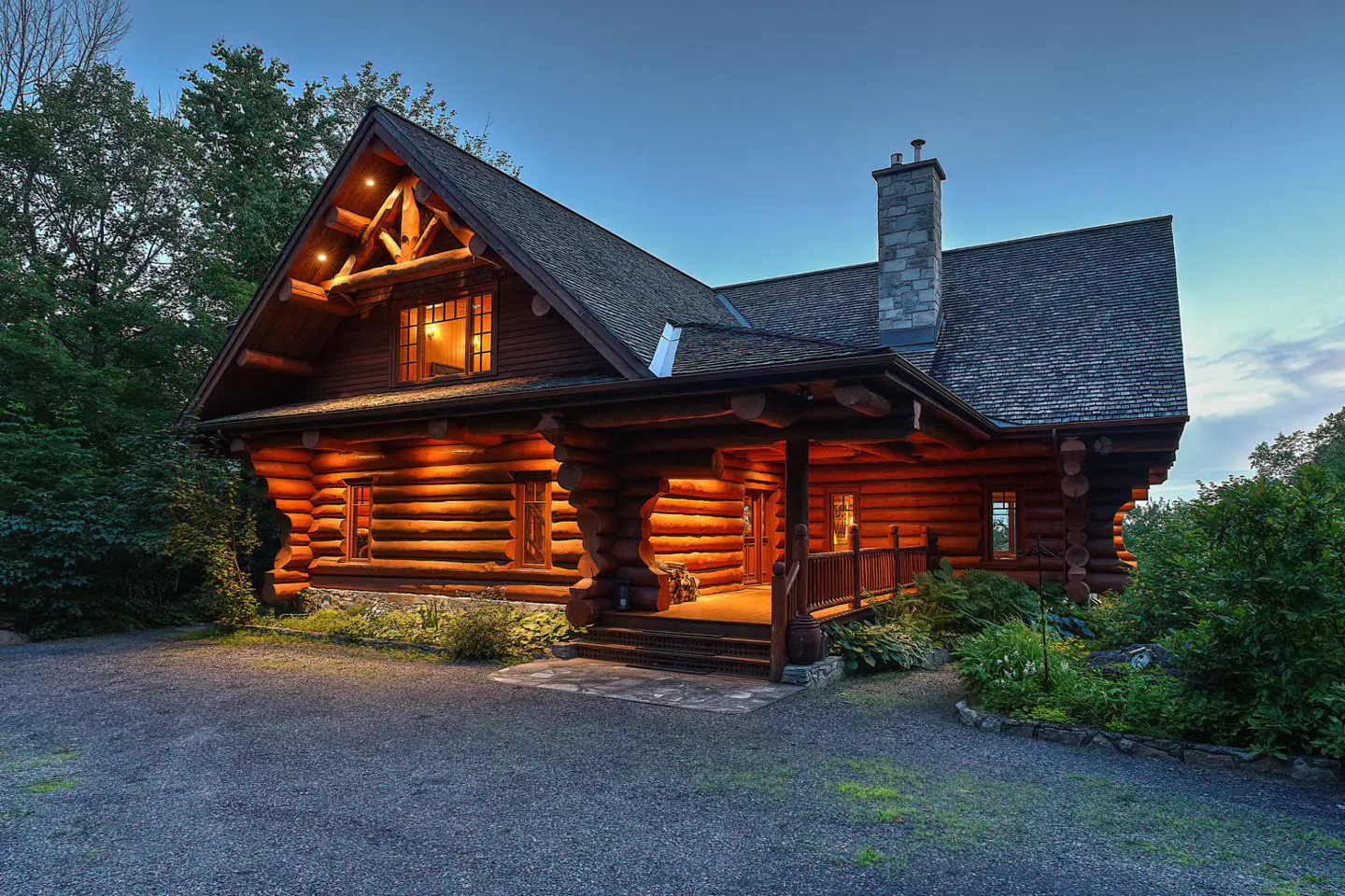 Exterior view of a log cabin home with a stone chimney and a covered porch at dusk.