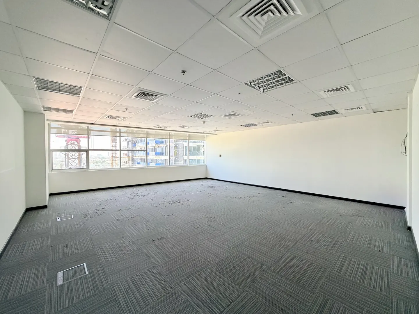 Empty office space with white walls, gray carpet tiles, a drop ceiling, and large windows overlooking a city construction.