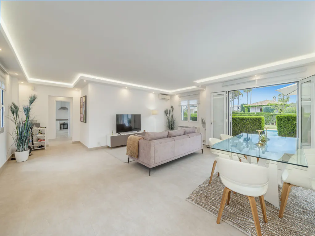 Bright, open-plan living and dining area with white walls, recessed lighting, and a large glass table with white chairs. A pink sofa faces a TV. Doors open to a garden.