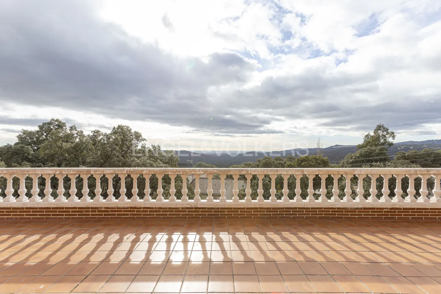Balcony view with terracotta tiles, brick base, and white balustrade overlooking trees and cloudy sky.