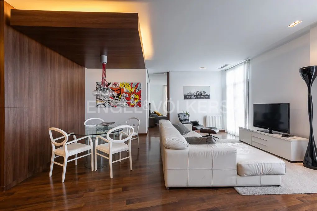 Open-concept living space with wood floors, white walls, and modern furniture. A glass table and white chairs are near a colorful art piece.