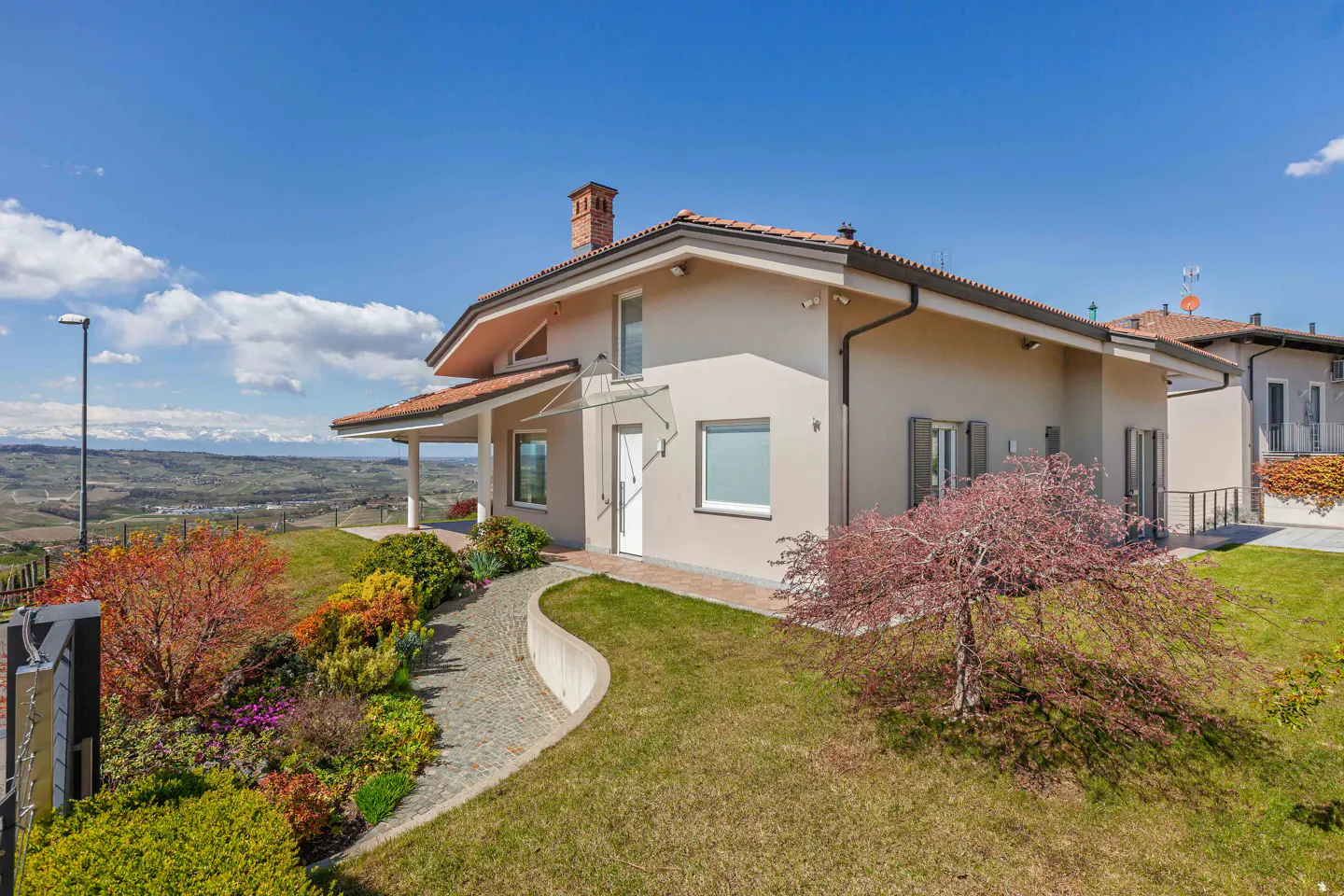 Beige two-story house with a red tile roof, green lawn, and colorful landscaping under a blue sky.