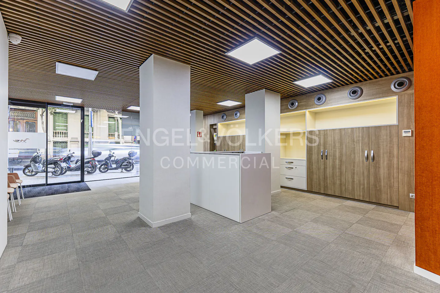 Real estate office interior with a white reception desk, wood cabinets, and a slatted wood ceiling. Motorbikes are visible through the glass entrance.