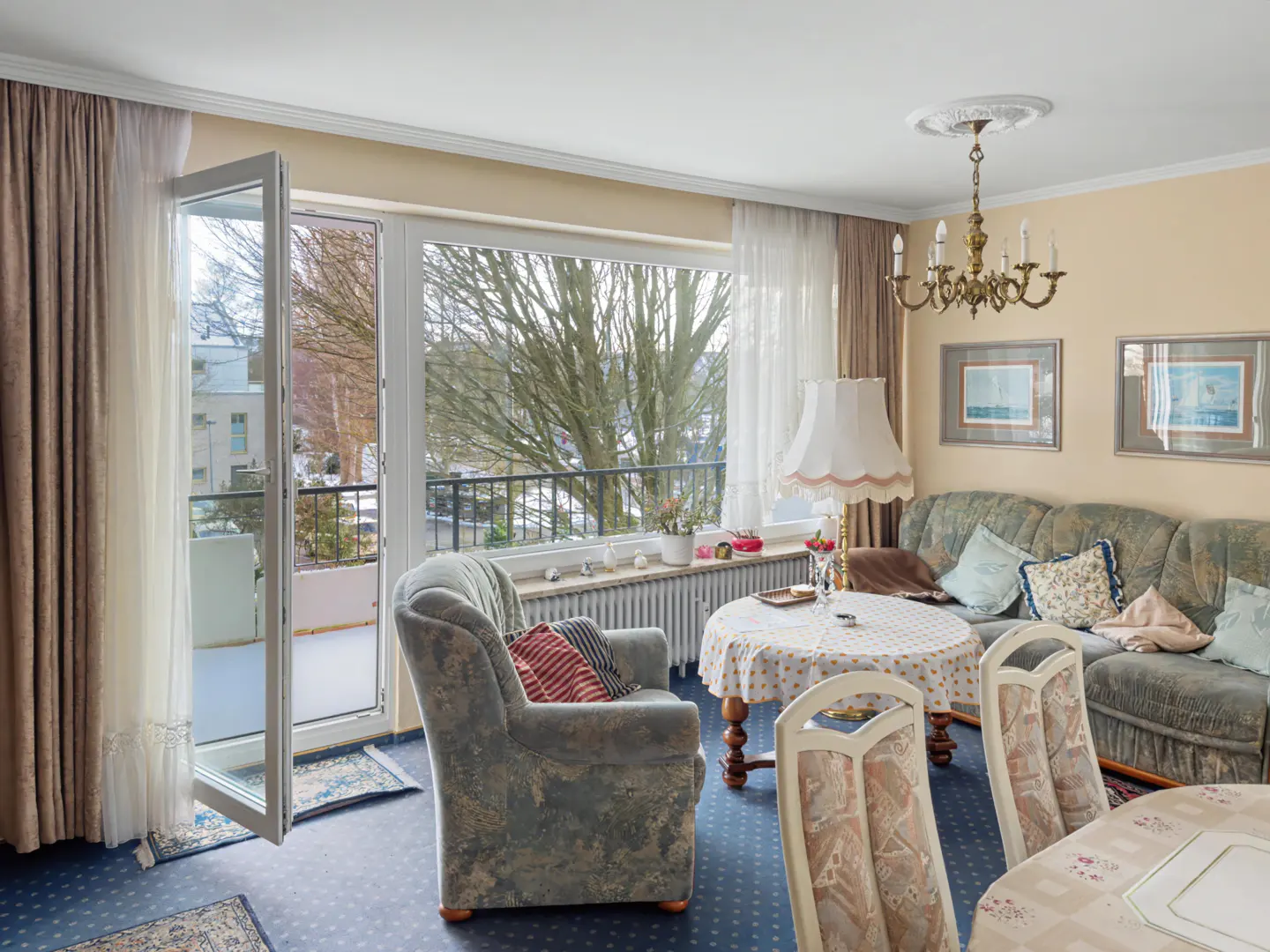 Living room with blue carpet, floral sofa and chair, round table, and open balcony door.