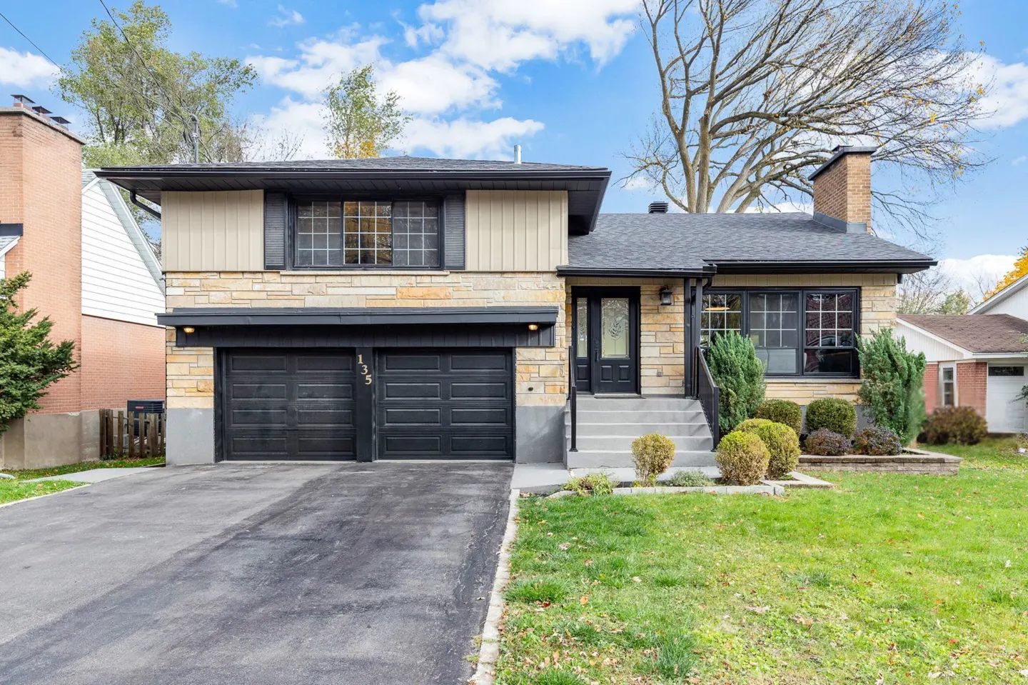 Two-story house with stone and beige siding, black trim, and a two-car garage. A lawn and trees surround the house.
