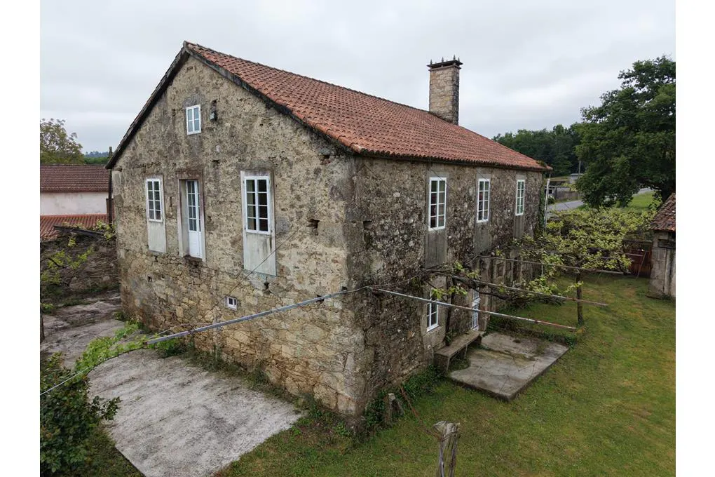 A rustic, two-story stone house with a terracotta tile roof, white-framed windows, and a stone chimney in a rural setting.