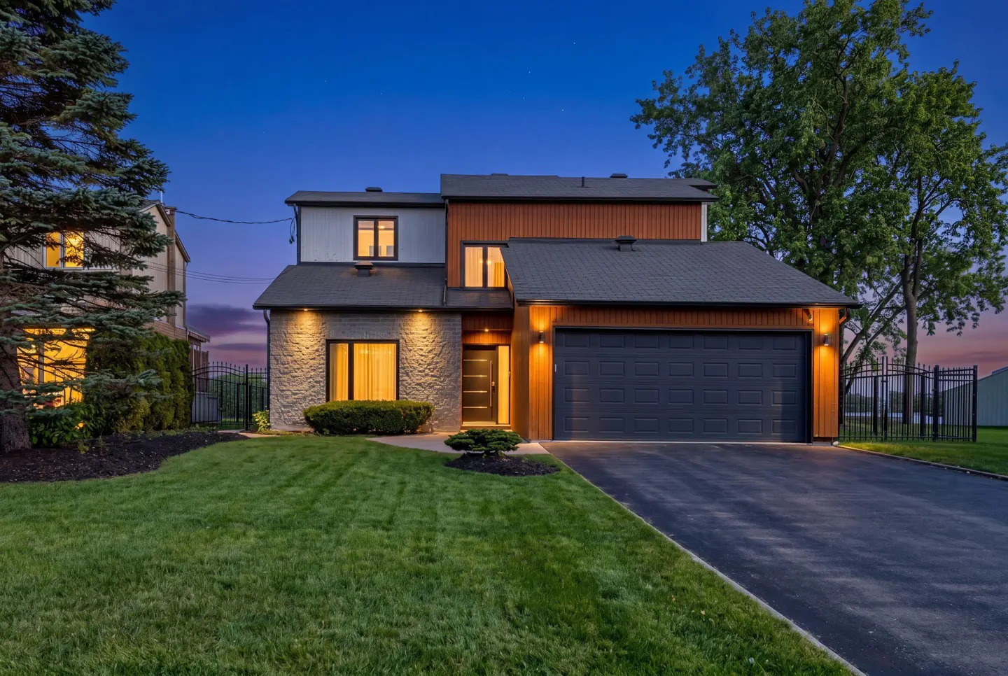 Modern two-story home with stone, wood, and white siding, a dark gray roof and garage door, and a green lawn at dusk.