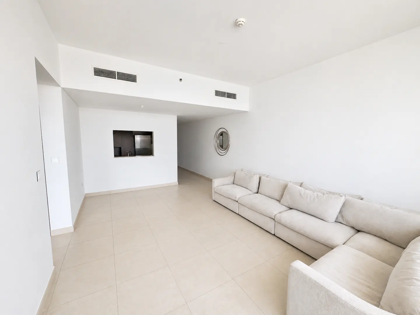 Bright, empty apartment living room with white walls, beige tile floor, and a large, light beige sectional sofa. A kitchen pass-through is visible.