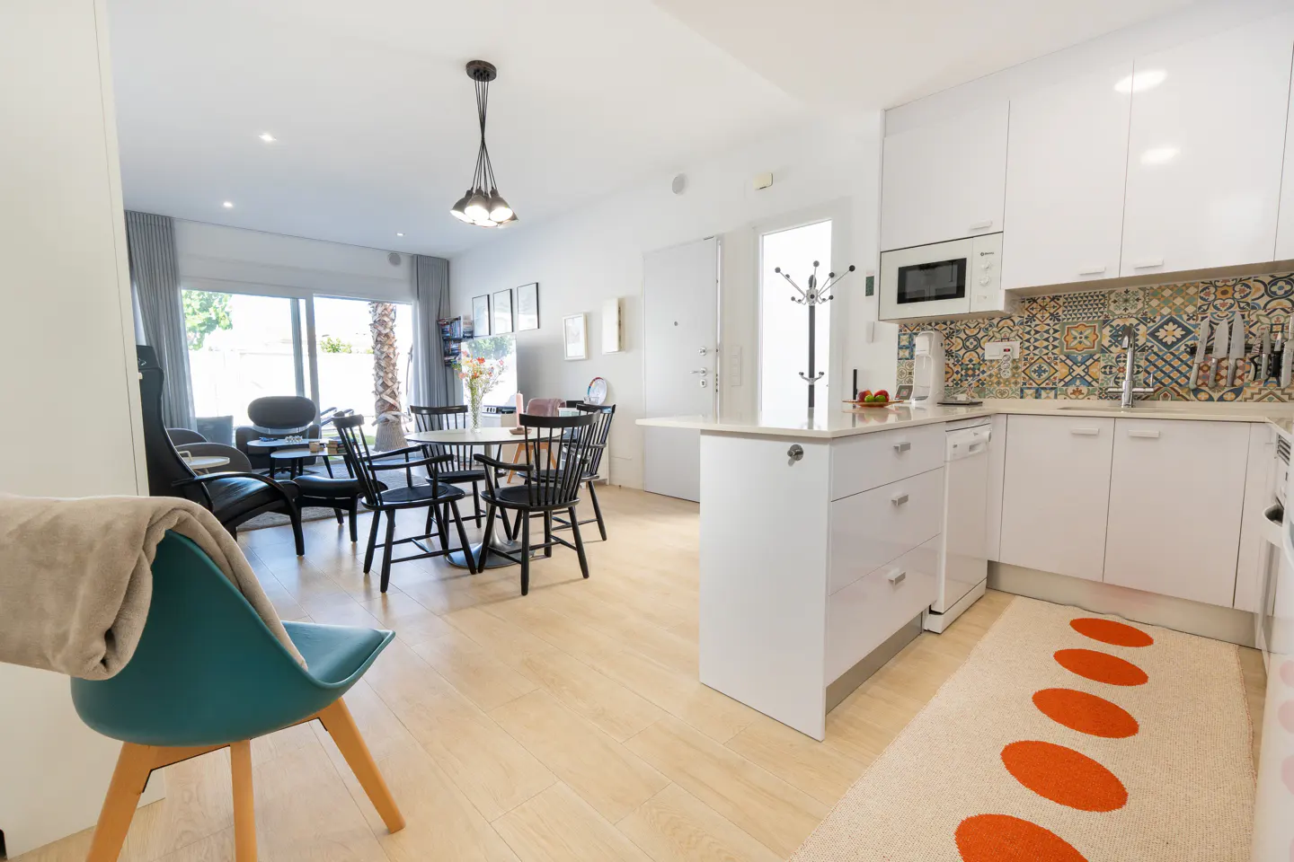 Modern open-plan kitchen and dining area with white cabinets, patterned backsplash, and a teal chair in the foreground.