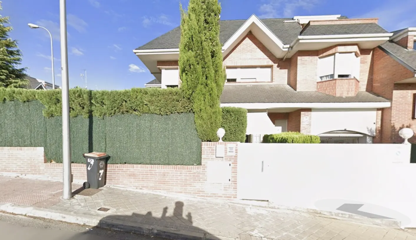 Two-story brick house with a gray roof, white trim, and a white gate. A tall green hedge and tree provide privacy. A trash can sits on the sidewalk.