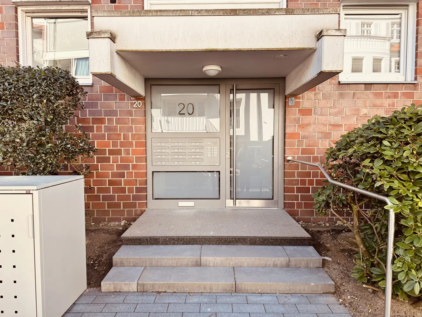 Exterior view of a brick apartment building entrance with a gray door, number 20, and steps leading up.