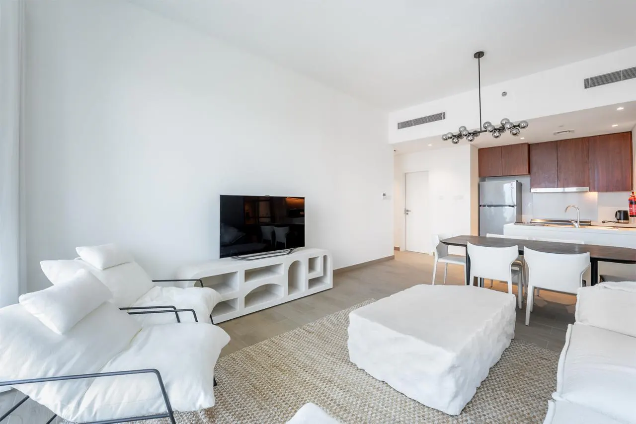 Bright, modern apartment living room with white walls, furniture, and a TV. Kitchen with brown cabinets in the background.