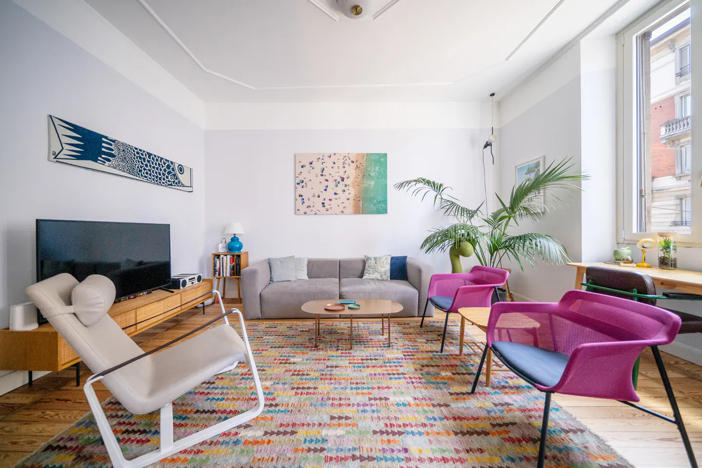 Bright living room with a gray sofa, pink chairs, and a colorful rug. A TV and artwork adorn the white walls. Natural light streams through a window.