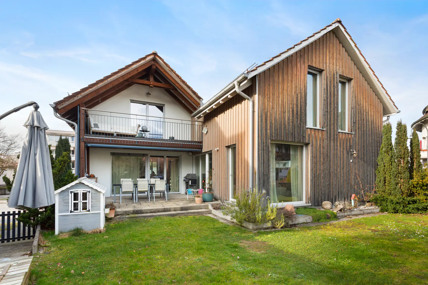 Two-story house with a wood-paneled addition, a patio with a table and chairs, and a green lawn.