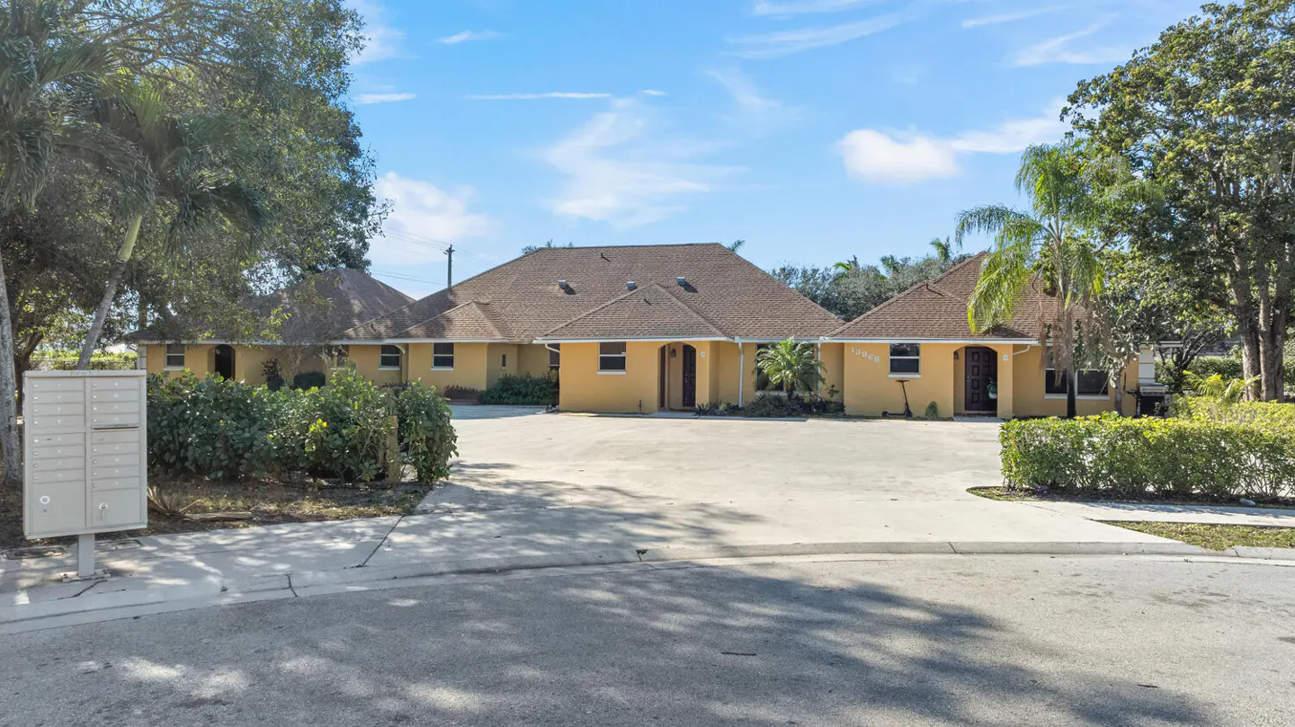 Exterior view of a yellow, one-story building with a brown roof and a concrete parking lot. Mailboxes and green bushes are in the foreground.