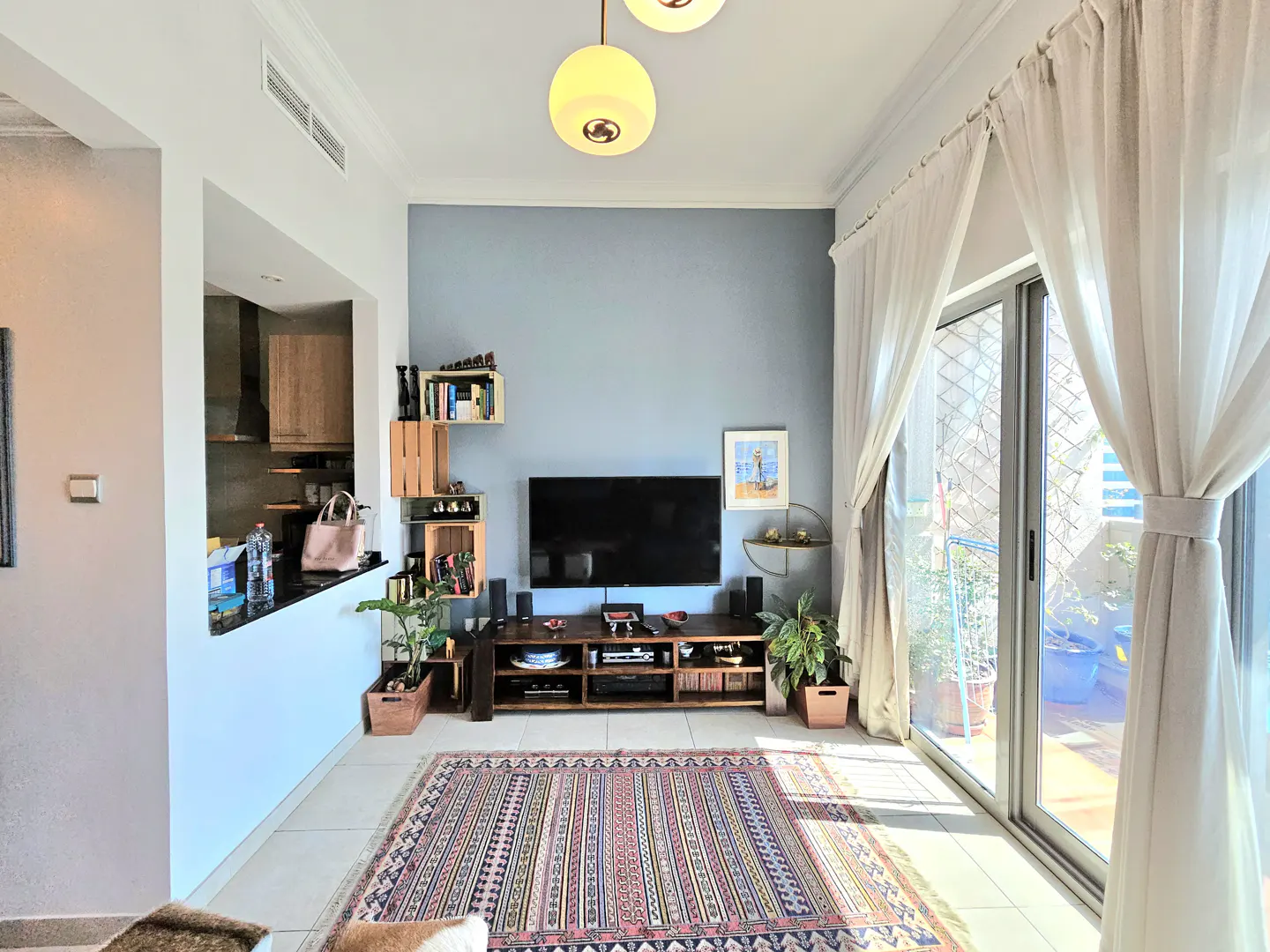 Living room with a TV, wooden shelves, and a patterned rug. White curtains frame a sliding glass door to a balcony.