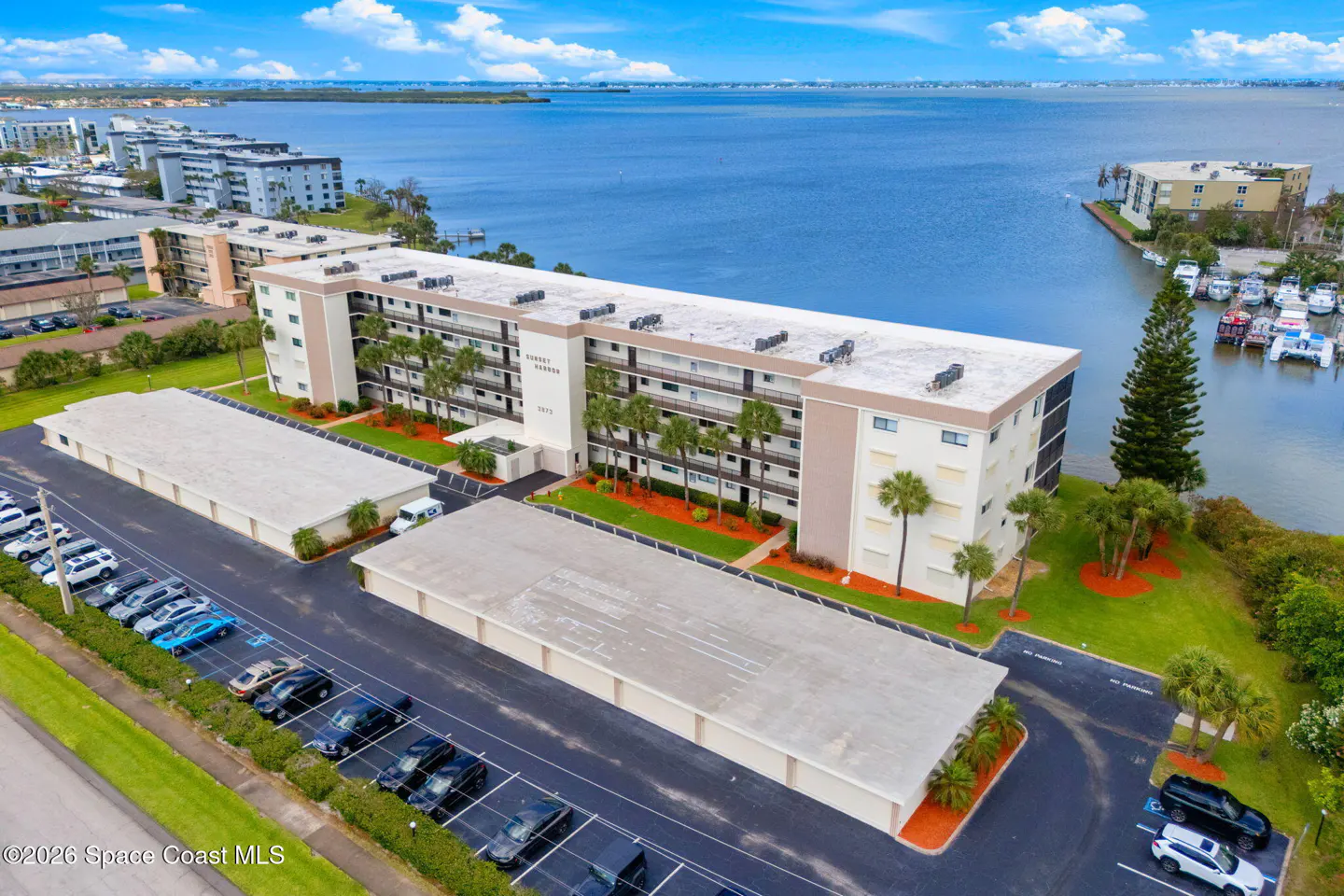 Aerial view of a beige condo building with palm trees, parking, and waterfront.