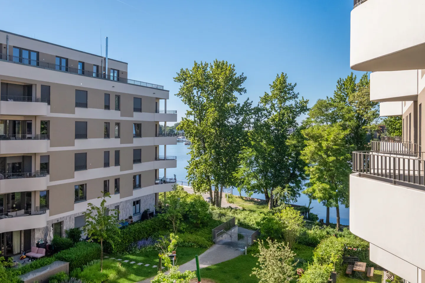 View from a balcony overlooking a modern apartment complex, lush green trees, a walking path, and a calm river beyond.