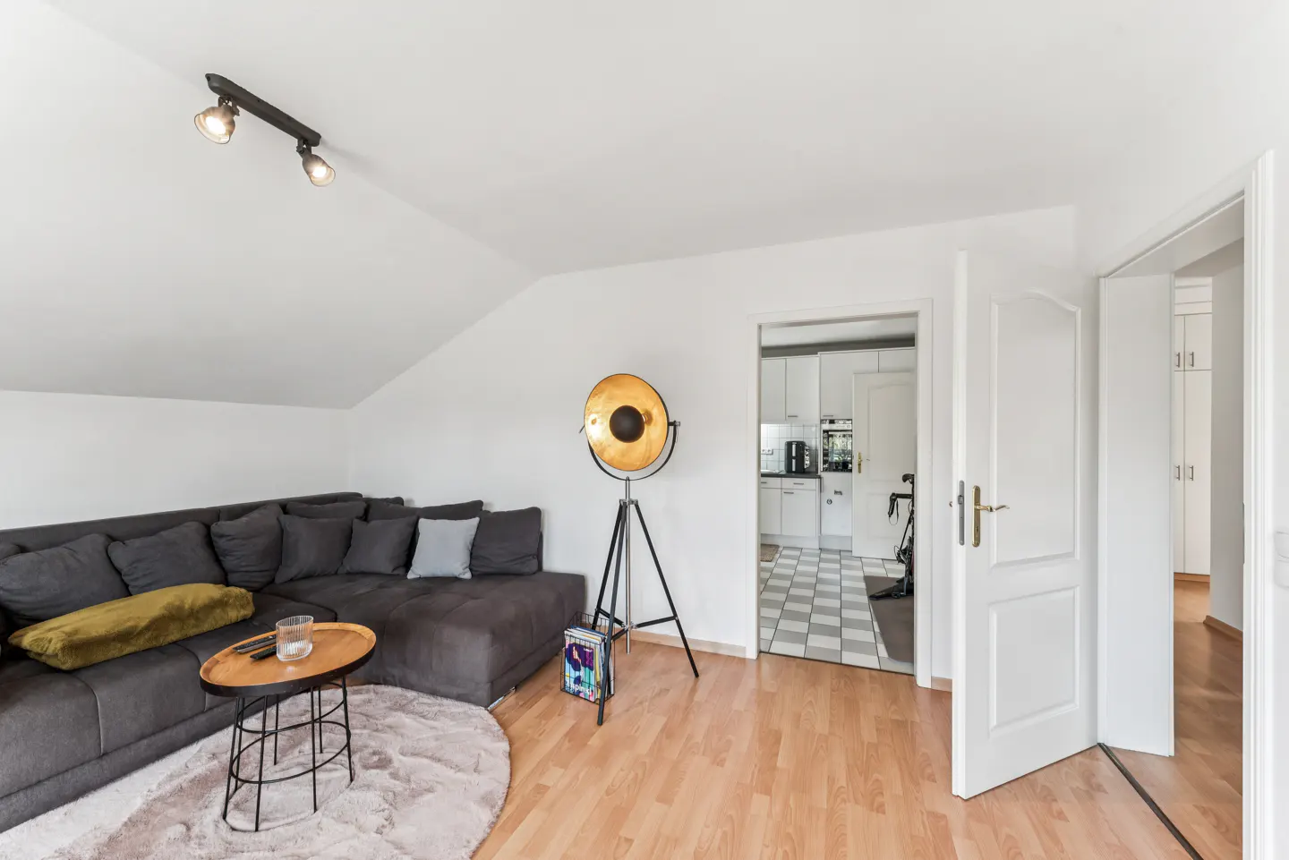 Living room with gray sectional sofa, round rug, and gold floor lamp. Kitchen visible through doorway. Hardwood floors and white walls.