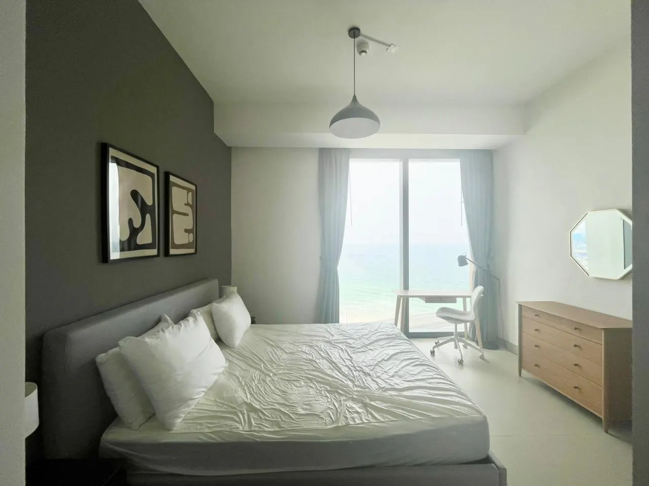 Bedroom with a gray bed, white linens, and pillows. A desk faces a window with ocean views. Dresser and abstract art add to the modern decor.