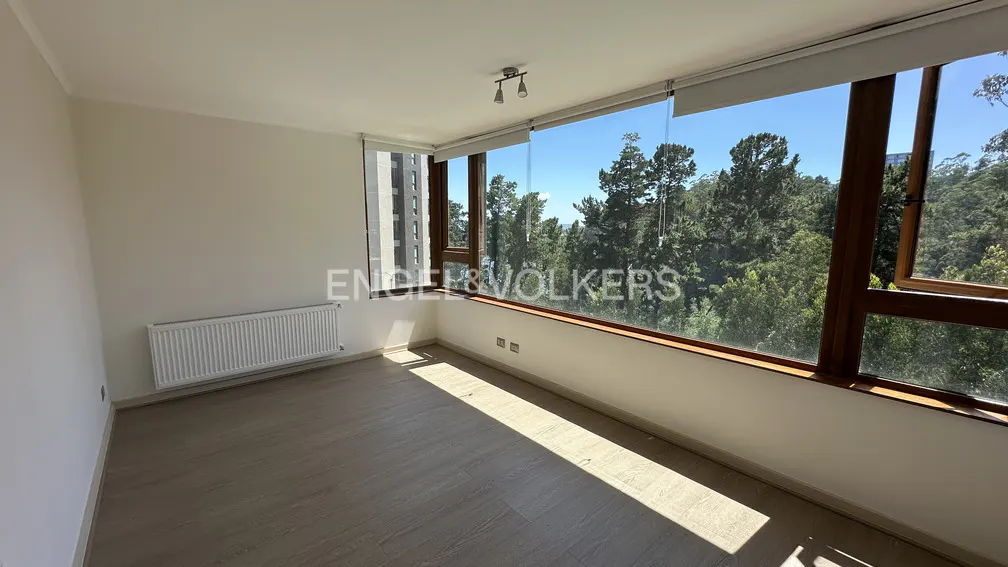 Empty room with light wood floors, white walls, a wall heater, and large windows overlooking a lush green forest.