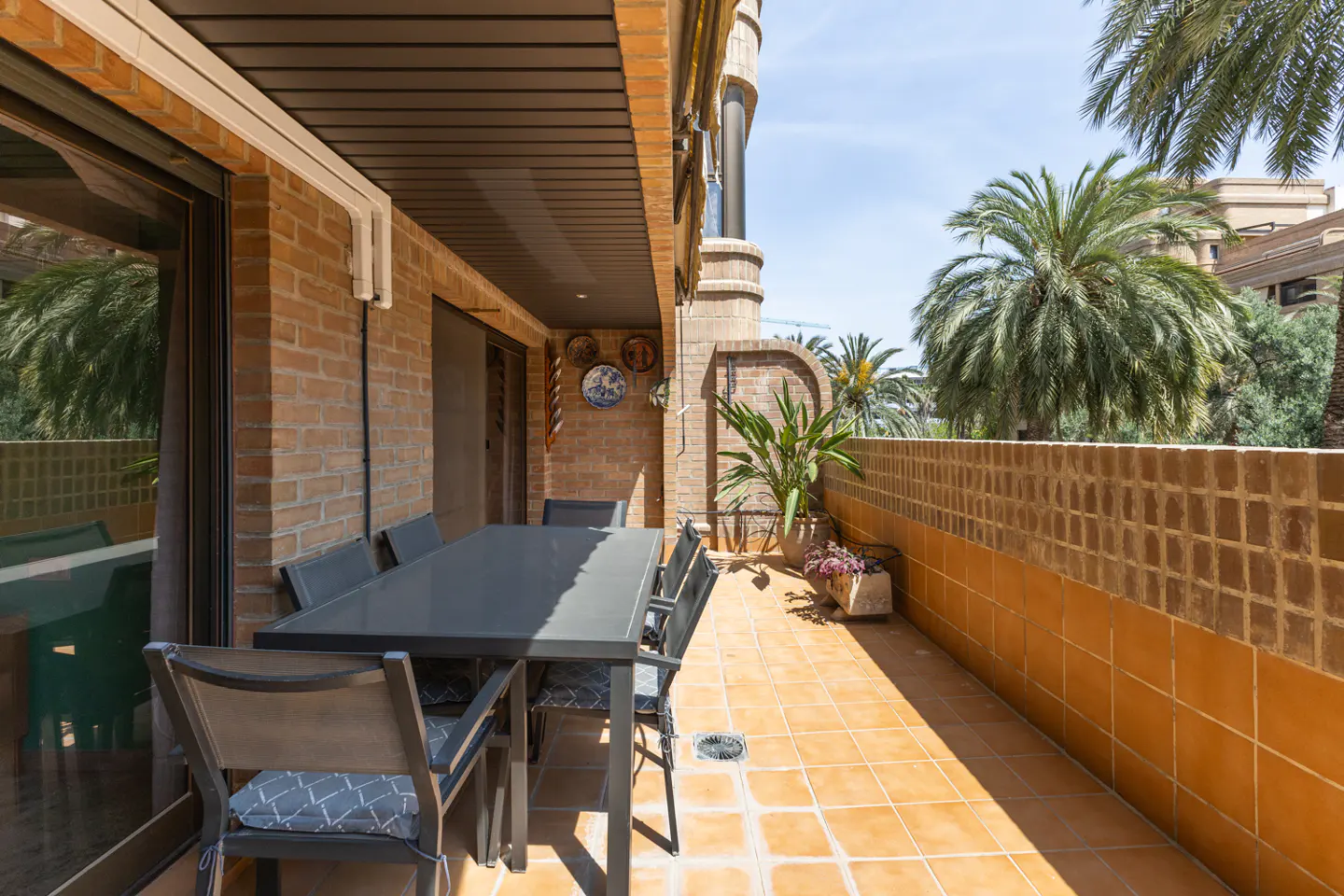 A sunny balcony with a long gray table and chairs, brick walls, and palm trees in the background.