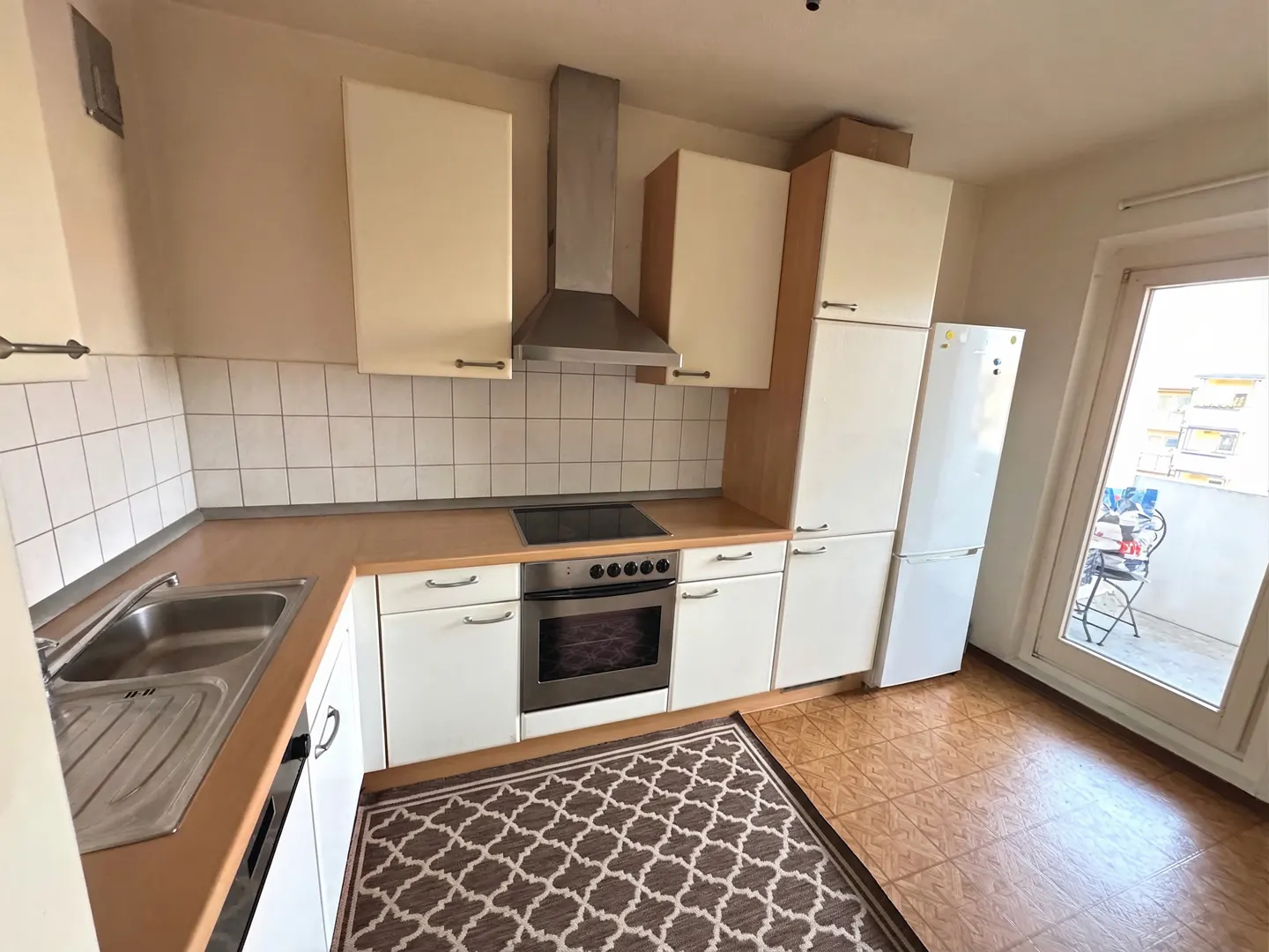A bright kitchen with white cabinets, stainless steel appliances, and a brown patterned rug. A glass door leads to a balcony.
