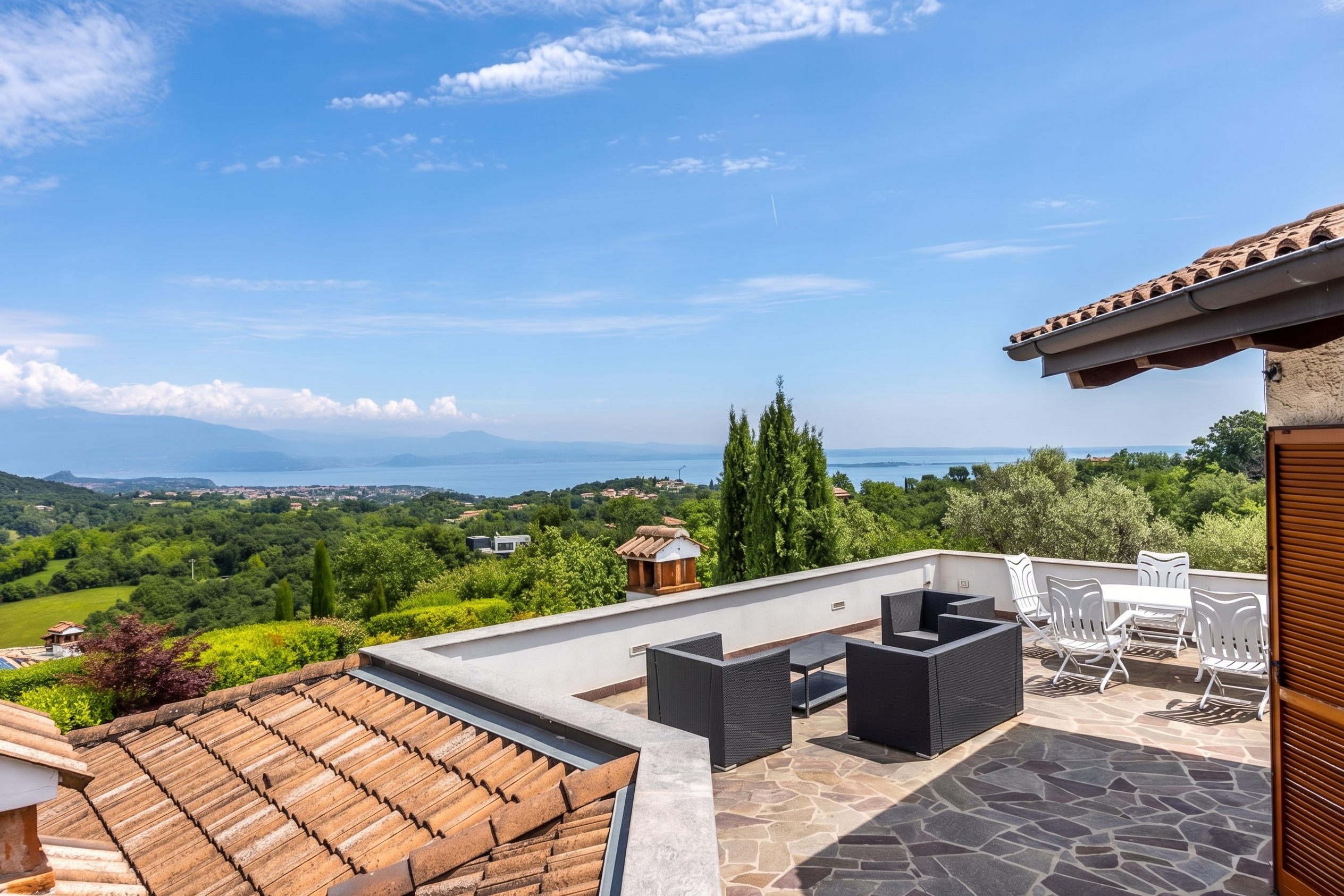 Outdoor patio with black wicker furniture and white table set overlooking green hills and a lake under a blue sky.