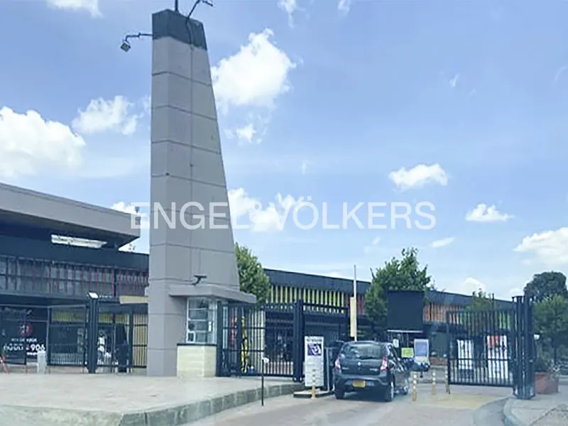 Exterior view of a gated property with a tall gray tower and a black car entering. Blue sky with clouds. Engel & Völkers logo overlay.