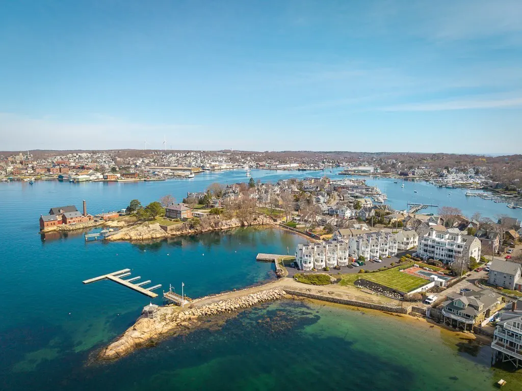Aerial view of coastal real estate with blue water, docks, and a clear sky. Buildings line the shore, showcasing waterfront living.