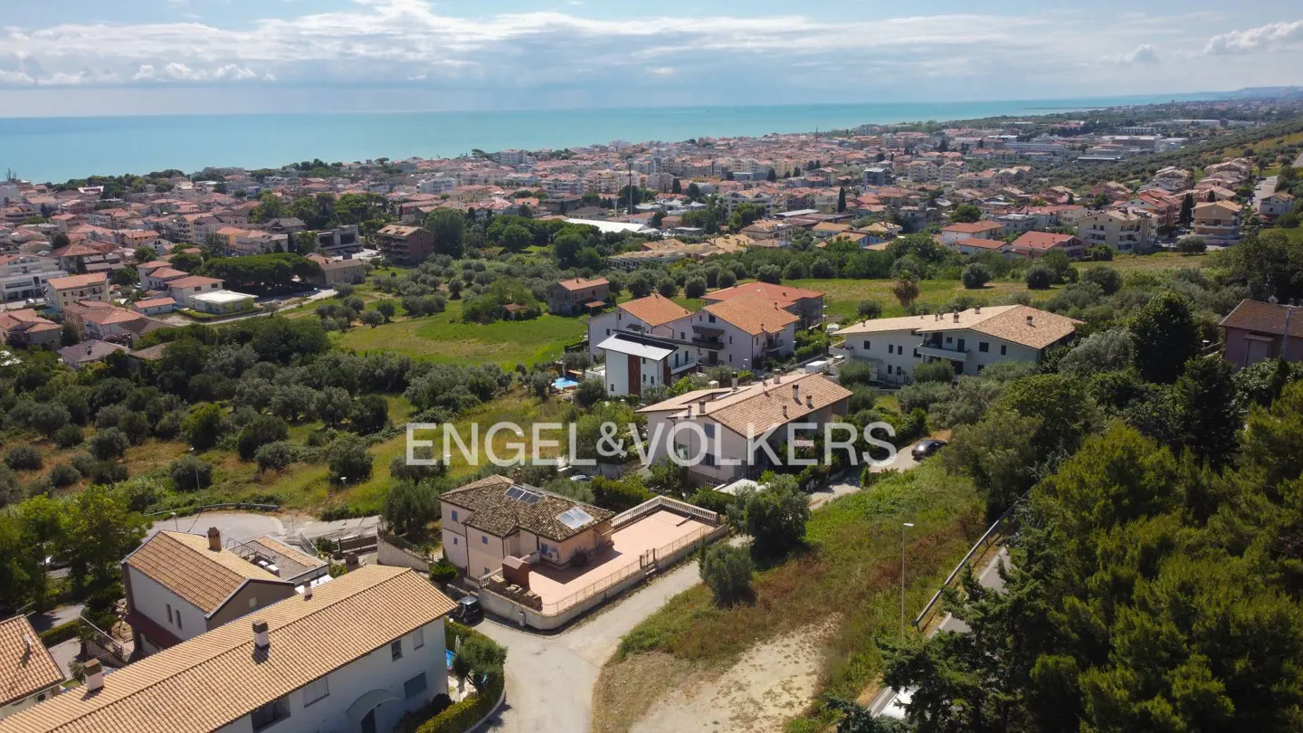 Aerial view of houses with red tile roofs, green trees, and the blue ocean in the background. Engel & Volkers logo is visible.