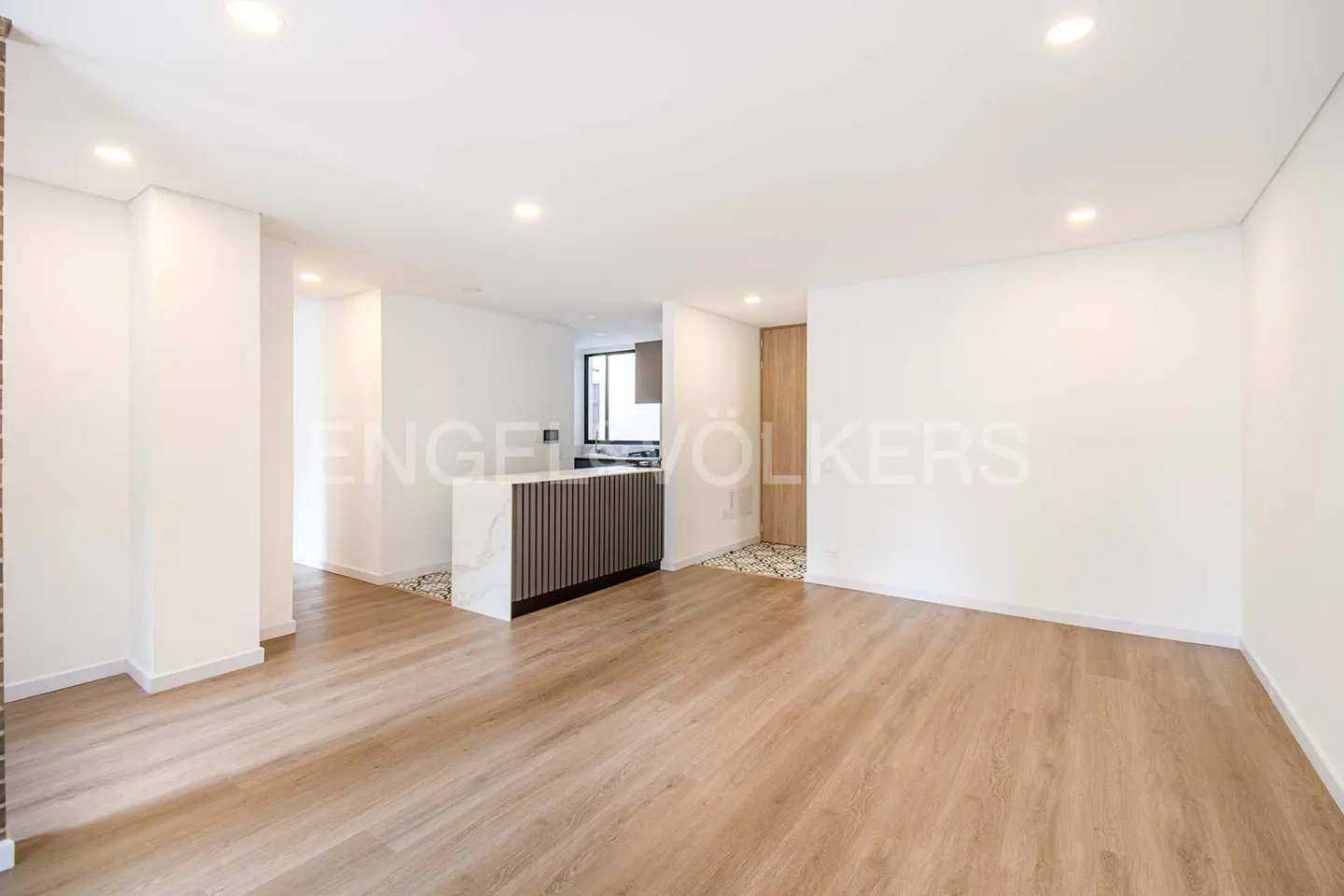 Bright, modern open-concept living space with wood floors, white walls, and a kitchen island. Engel & Völkers logo visible.