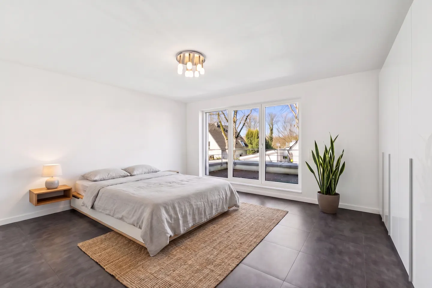 Bright bedroom with white walls, dark tile floor, and a large window. A bed with gray linens sits on a jute rug. A potted plant stands near a white wardrobe.