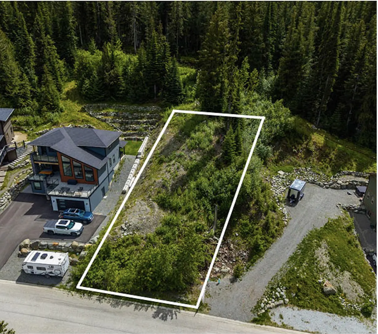 Aerial view of a vacant, wooded lot outlined in white, situated next to a modern house and a gravel driveway.