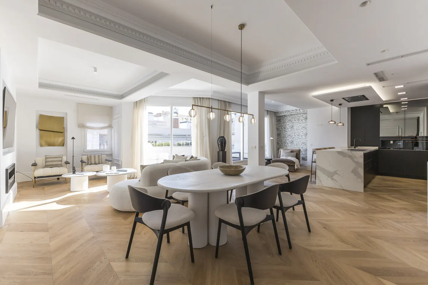 Open-concept living space with herringbone wood floors, white walls, and modern furniture. Dining table with black chairs and kitchen island visible.