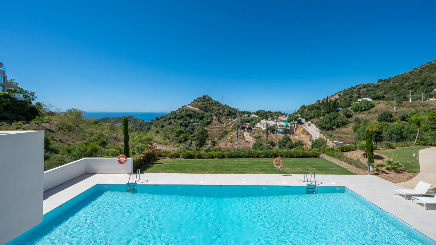 A turquoise pool with ladders sits before a green lawn and tree-covered hills under a clear blue sky.